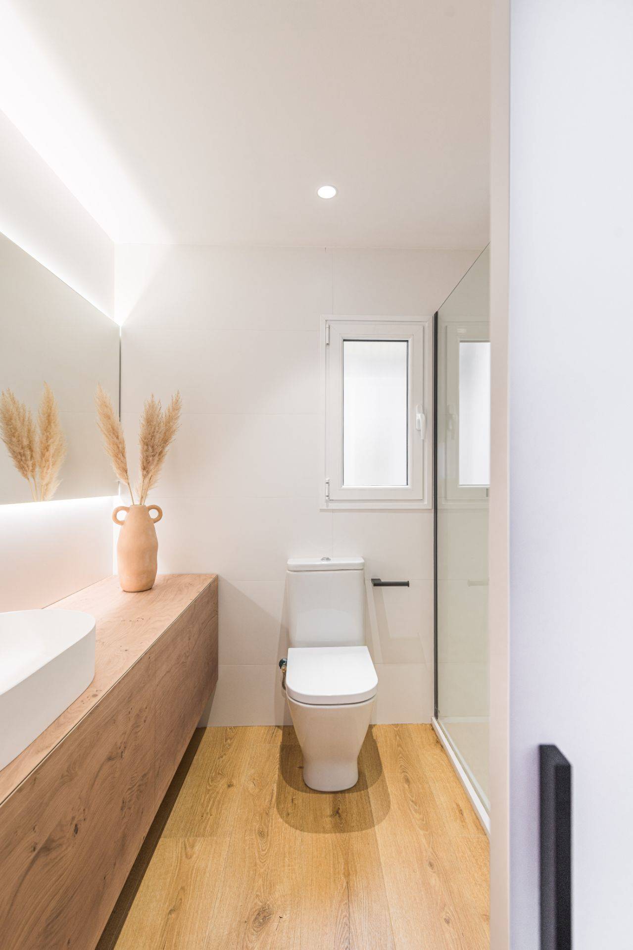 Baño moderno después de la reforma con ducha con mampara, suelo de madera, mueble del baño de pared a pared y espejo iluminado. 